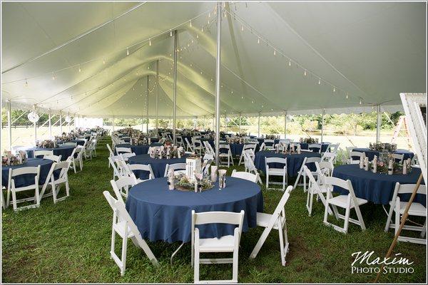 Wedding Tent Rental in Cincinnati, with bistro lighting, white padded garden chairs, round guests tables, navy linens and tent pole drapes.