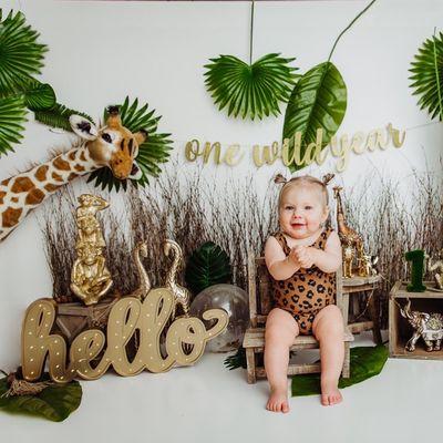 Wild One baby girl in a photo shoot with grasses and large stuffed giraffe and leaves.