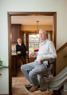 man riding curved stair lift with wife in background in kitchen