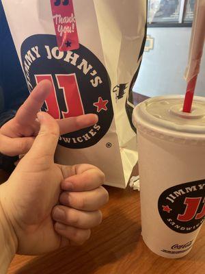 two hands displaying a peace sign and thumbs up in front of a jimmy johns bag and drink cup