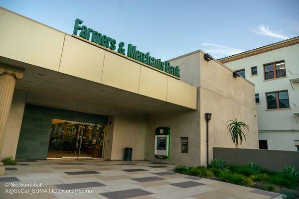 FRONT OF BUSINESS: The entrance at evening. A large sign reads, "Farmers & Merchants Bank" and an ATM is located underneath the porch roof.