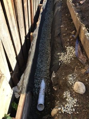 New French Drain installed behind a retaining wall. The trench is wrapped with a fabric and the pipes are buried under drain rock.