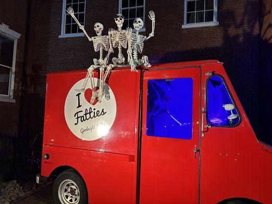 Picture does a red delivery van with three skeleton to send it on top and a sign on it. That says I love Fattys.