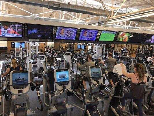 Full rows of cardio machines along the wall facing the weights and TVs