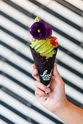 Matcha soft serve in a midnight vanilla cone topped with strawberries , pistachio, edible flower , & dusted with matcha  powder.