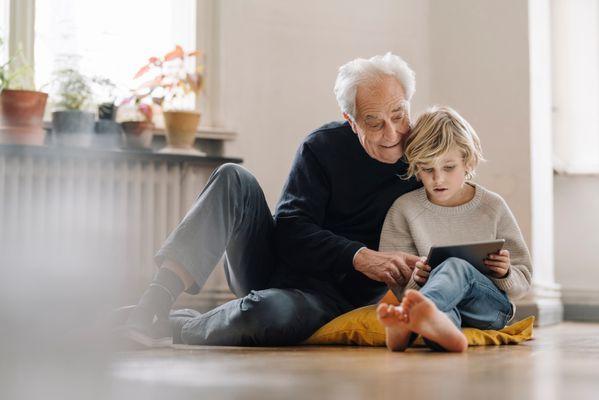 An elderly man spending time with his grandson on his tablet while sitting on a cushion.