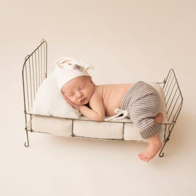 Newborn boy Jonah in newutral cream color sleeping on metal bed with a sleepy hat and stiped brown pants