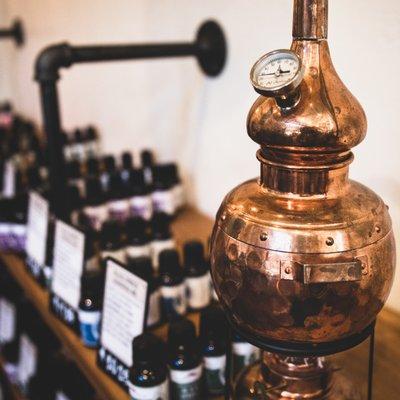 A small copper essential oil still surrounded by colorful essential oil bottles.