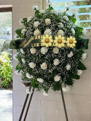 Stand spray with white carnation and a few roses.  Wording in gold on a green banner/ribbon.