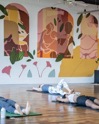 Students take a yoga class in the studio. The mural by Moon Juice Murals and Silent Fox Studio is in the background.