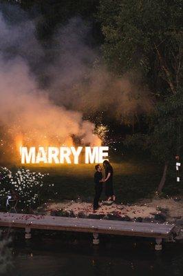 LeeReadian couple getting engaged in front of a giant, lit up marry me sign