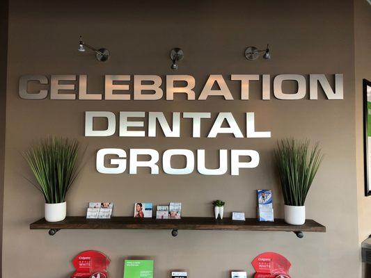 Interior wall sign of Celebration Dental Group with plants on either side of a brown wooden shelf.