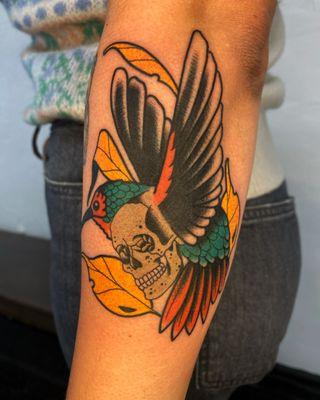 Neo traditional hummingbird/skull morph with gold leaves on the outer forearm. Make by Blake Aiken.