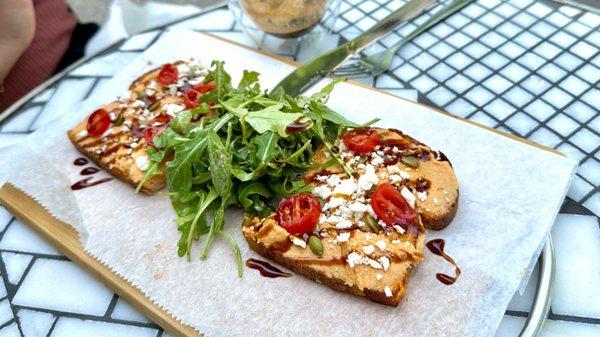 Hummus Toast topped feta, cherry tomatoes, and sunflower seeds drizzled with balsamic glaze and a side of arugula.