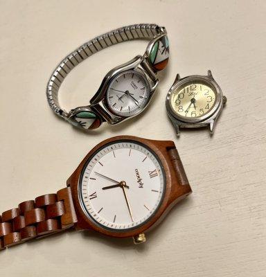 An aerial view of two watches and a watch face laying on a white table. One of the watches is wooden. The other is silver.