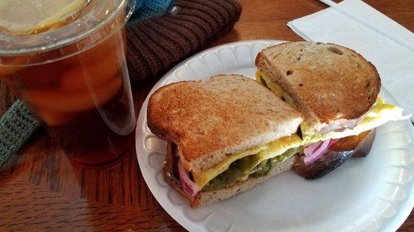 An omelet sandwich with red onion,  tomato,  and green chile on toasted multigrain bread, with an iced tea.