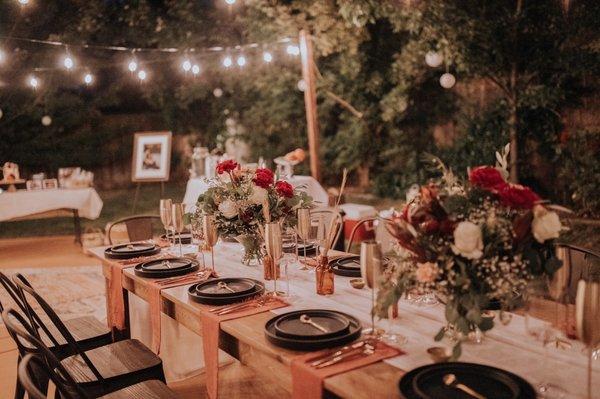 Farm house table, black meta chairs, ceramic plates, drink ware, utensils, linen, and string lights.