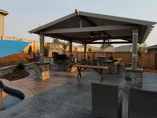 Freestanding patio cover with a pan roof, covering a built in kitchen and bbq. Equipped with fans and outlets. Finished with stone pillars.