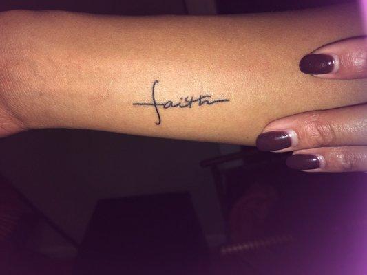 The tattoo says "Faith" in cursive and also in the shape of a cross.