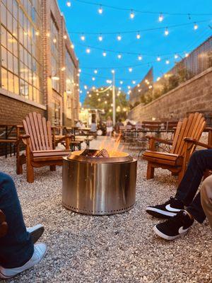 Fire pit with flames coming out of the top and string lights against a pale blue night sky