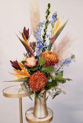 Autumn arrangement in a gold vase with birds of paradise, cremones, roses. delphinium, leucodendron and pampas grass.