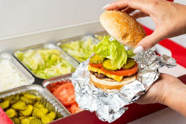 A close-up photo of a person putting the top bun of their cheeseburger in front of a Five Guys catering box of toppings.