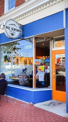 Storefront of The Grind, with the store sign over the large picture window, and the door to the right of the windows