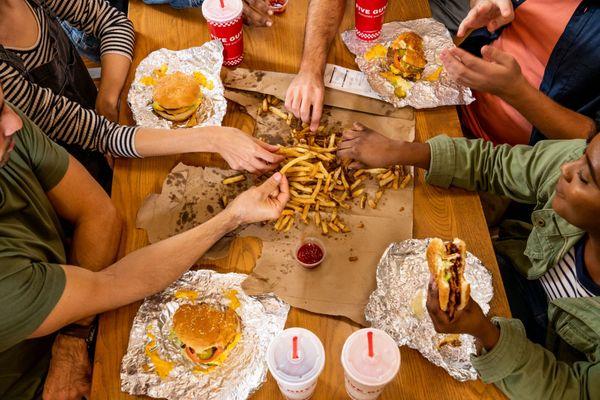 A top-down photograph of a group of friends sharing an order of fries alongside their sandwiches at a Five Guys restaurant.