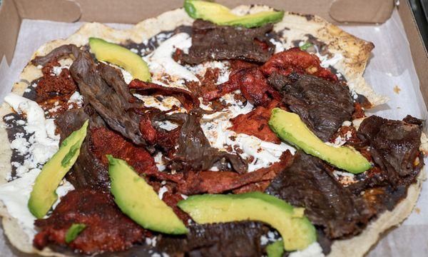 Tlayuda is a large tortilla spread with refried beans and topped with avocado, various meats, Oaxaca and fresh cheese.