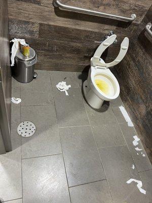 Super gross bathroom with sticky, smelly floor & broken toilet seat to accentuate the trash and pee all around.