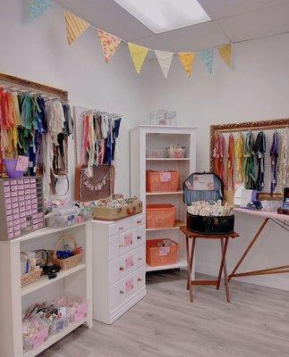 Corner of a store with brightly colored zippers hanging from frames. White furniture with baskets of products on on the shelves.