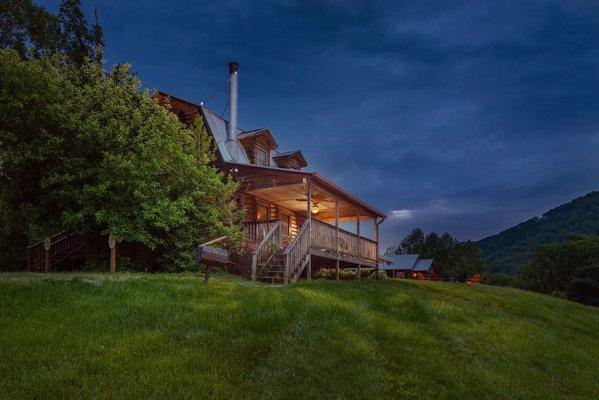 Exterior of the Mt. Mitchell cabin in the evening, showing warm cabin lights and a sweeping view of the mountains