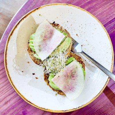 Avocado Toast with lemon, chili flakes, sea salt, alfalfa sprouts, and radish on a sprouted seed and grain muffin