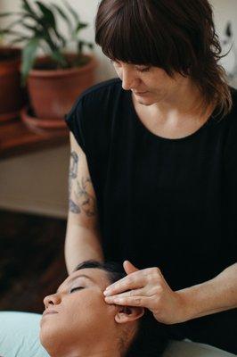 A practitioner wears a black top and gently works on a clients face. Blue sheets can be seen under the client and plants in the background.