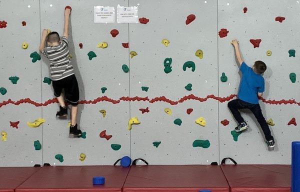 2 boys climbing the YMCA climbing wall while doing their obstacle course. An 8 yr old and 11 year old.