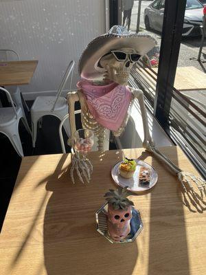 A skeleton dressed up in a hat and glasses enjoying a treat at a table in Blondie's