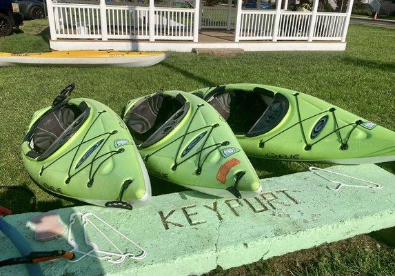 Three kayaks on a lawn near a gazebo propped up on a long green rectangle of concrete with the word "Keyport."