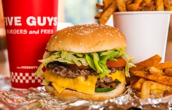 A close-up photograph of a Five Guys cheeseburger, soft drink in red Five Guys cup, and regular order of fries sitting on a t...