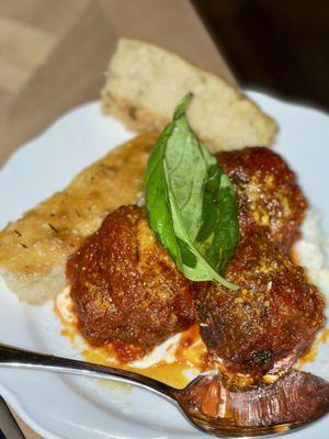 Delicious meatballs (beef/pork) with marinara sauce, melted cheese, a basil garnish, and a slice of herbed bread.