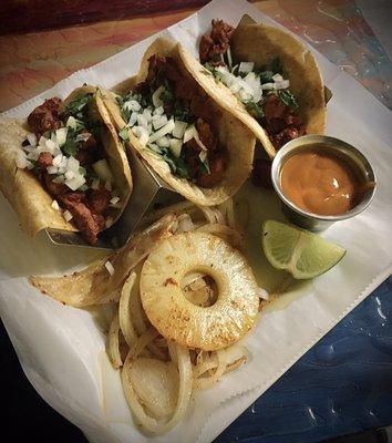 Tacos al pastor (pork) with cooked onions, a grilled pineapple slice, and mild chipotle sauce.