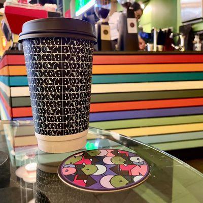 Hot coffee cup and coaster in Brooklyn Roasting Company's repeating logo design on table with barista counter in background.