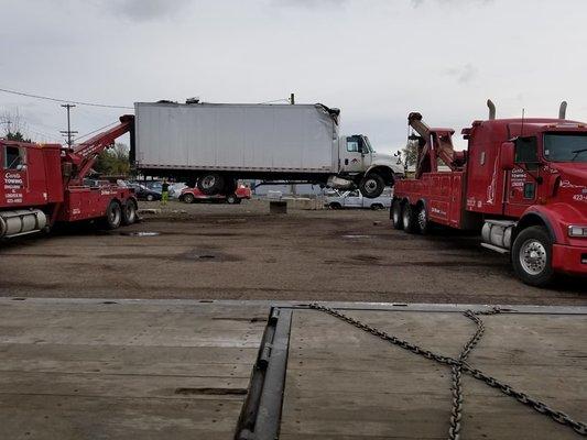 The men to know when you need a tow. Two trucks lifting a large box truck that has damaged wheels.