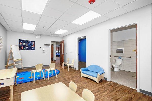 Classroom with brightly colored rug and whiteboard. Bathroom is seen through a door.