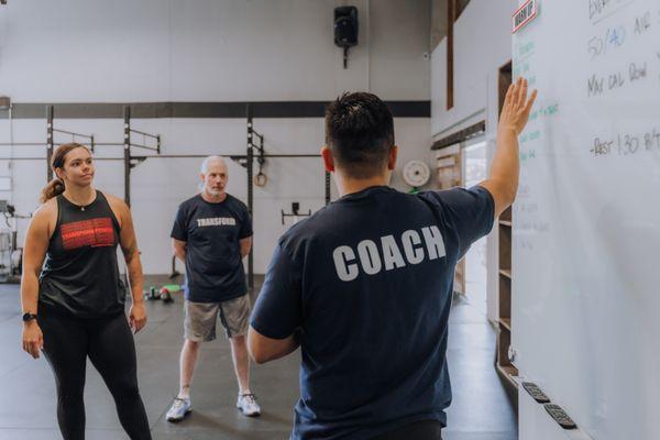 "A coach explains the workout to members, who listen intently. Equipment and racks are in the background."