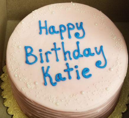 A round, gluten-free birthday cake with pale pink icing & white edible "pearls." The words "Happy Birthday Katie" are in the middle in blue.