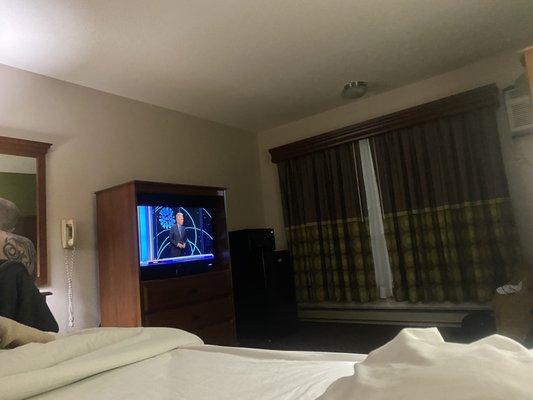 Bed, TV, big window, mini refrigerator and microwave and part of a brown chair in the corner