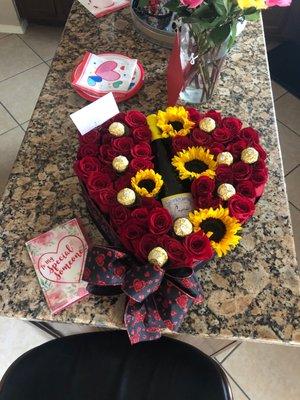 A roses and sunflower filled heart shape pot with wine bottle and chocolates.