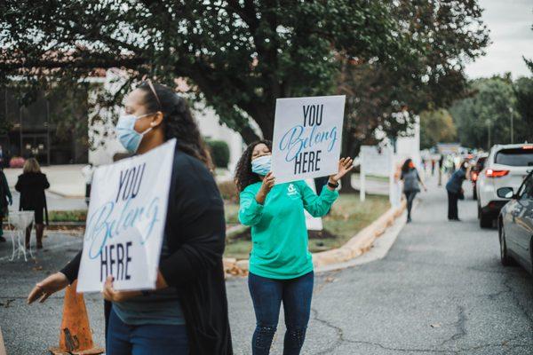 A young lady welcoming incoming cars with a hand held sign that reads, "You Belong Here."