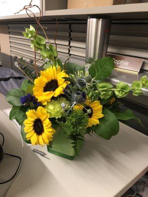 Flower arrangement sitting on a desk.  Primarily sunflowers and greenery, in a cute square glass vase.