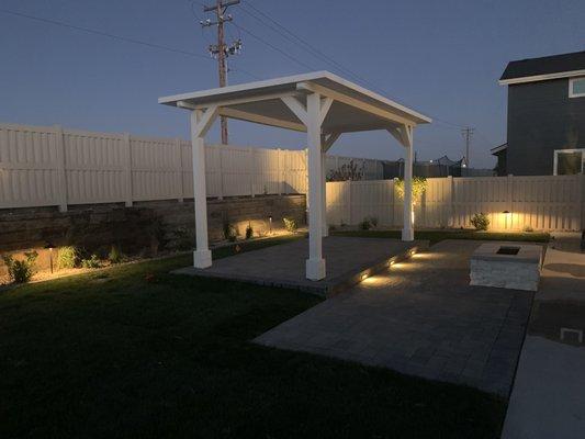 10x18 raised Pergola on pavers with lights illuminating the step. Fire pit and LV lights